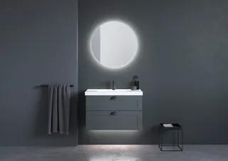 Svart badrum med rund spegel, grå handduk på stång, svart handfat med skåp, svart pall med handduk.